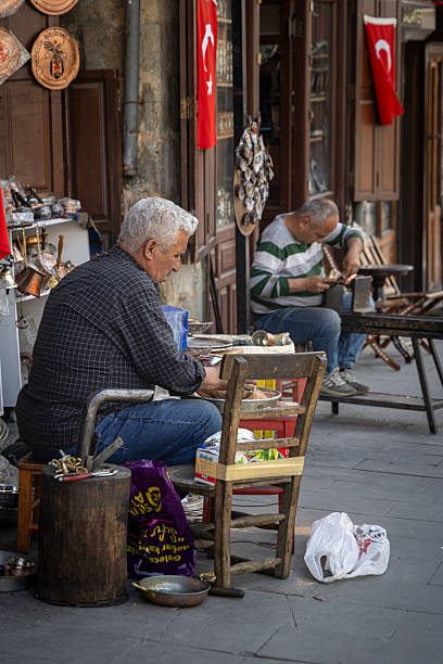 Istanbul, Turkey - April 18, 2025: A traditional copper craftsman working outside his shop in the historic bazaar district of Gaziantep, Turkey. The artisan is shaping a piece of copper using hand tools, a practice that has been passed down through generations. Surrounding the workspace are shelves filled with handmade copper goods, souvenirs, and local crafts. A Turkish flag is displayed above the shop, highlighting the cultural and national pride associated with traditional craftsmanship in southeastern Anatolia.
