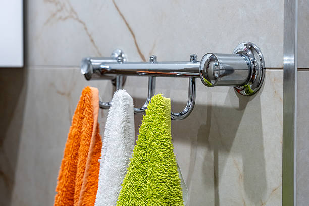 Soft microfiber towels in vibrant orange, white, and green colors hanging on sleek chrome hooks attached to a tiled bathroom wall, representing cleanliness and bathroom organization