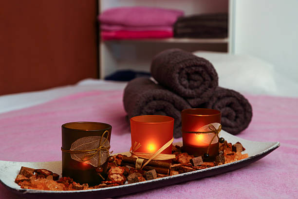 A tranquil close-up of rolled brown towels and lit candles on a decorative plate with aromatic elements, set on a light purple surface in a spa. The image evokes a sense of relaxation, wellness, and a calming spa atmosphere. Ideal for illustrating themes of spas, wellness, relaxation, aromatherapy, and self-care in Europe.
