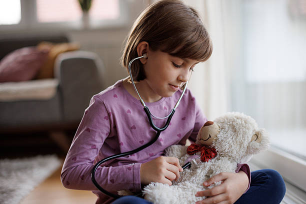 istock Cute little girl playing doctor with her teddy bear, using a stethoscope while sitting by the window at home 2212458594