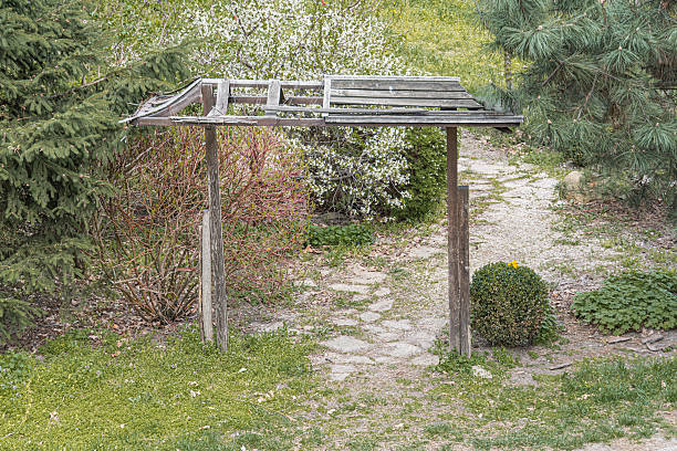 A weathered wooden pergola in a park setting, situated between trees and a stone path Muted colors and soft lighting suggest an overcast day