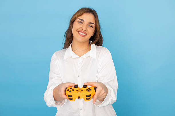 istock Young woman holding a video game controller, smiling at camera on blue background, enjoying her leisure time 2212460094