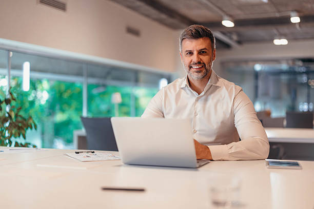 istock Businessman working with laptop and smiling at camera in modern office 2212461377