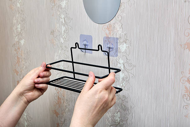 istock Hands are installing black metal bathroom shelf onto wall mounted transparent adhesive hooks by aligning hooks with designated attachment points. 2212461467
