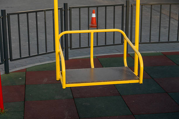 istock Bright yellow swing set stands empty in a park with colorful rubber flooring, surrounded by a black fence and a traffic cone nearby 2212462278