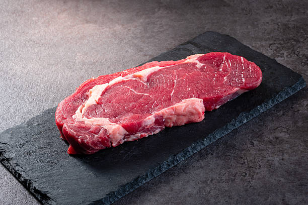 Raw beef steak with visible fat layers placed on a black slate board over dark textured background. Clean presentation for butcher shops, food styling, menu visuals, or cooking themes.