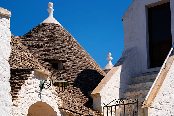 Close-up view of traditional Trulli rooftops with their iconic conical shapes, alongside white stone building in Alberobello, Italy. Bright blue sky contrasts with rustic architecture
