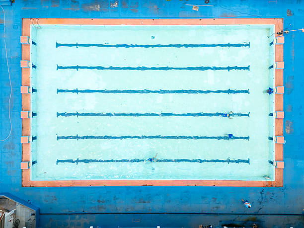 Drone top-down view of an olympic-size swimming pool with few swimmers practicing, capturing the clear blue water, swimming lanes, and athletic activity from above