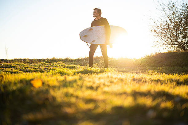 Man in a wetsuit carries surfboard through sunlit green meadow in spring, heading towards an unseen destination. Adventure concept.