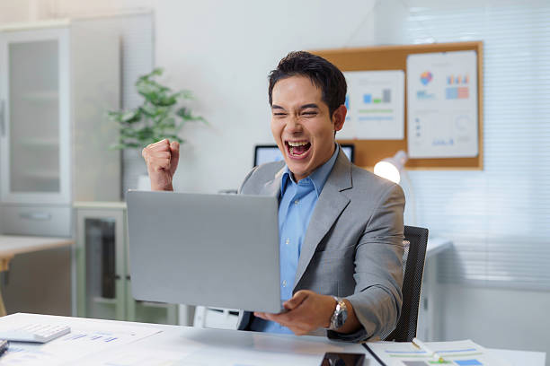 istock Young businessman expressing excitement and joy while looking at his laptop, celebrating a successful business achievement in his modern office 2212497533