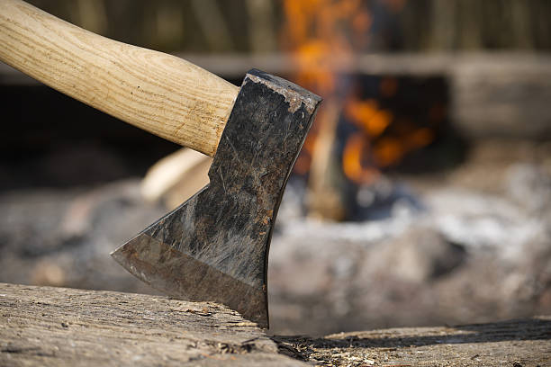 istock An axe is deeply embedded in a wood chopping block with a warm campfire blazing behind it. 2212505957