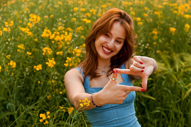 Young woman in denim dress and jewelry posing playfully among blooming yellow flowers on a sunny day