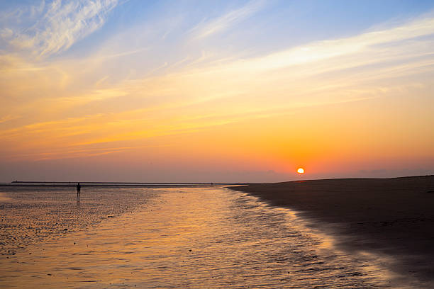 A peaceful beach landscape at Isla Canela in Huelva, Spain, during a vivid sunset. Warm tones of orange and yellow fill the sky, reflecting softly on the wet sand and calm shore, creating a tranquil and scenic coastal view.