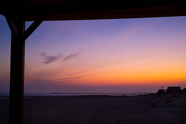 View of the vibrant evening sky from under a canopy at Isla Canela beach in Huelva, Spain. The soft gradient of orange to purple captures the beauty of dusk, creating a peaceful and scenic coastal moment with silhouetted elements in the foreground.