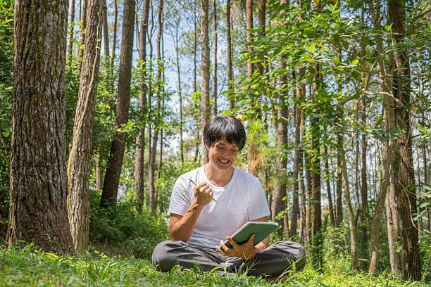 istock A young asian man wearing a white t-shirt and gray pants is drawing using a digital tablet while sitting on the grass in a lush pine forest in the morning. Remote work concept 2212516741