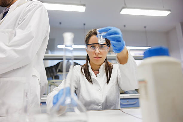 Female university student wearing lab coat, surgical gloves and protective eyewear for research with chemicals in practice class in the lab with colleagues
