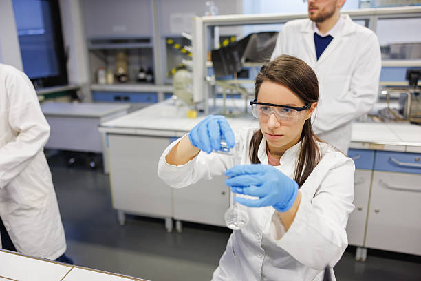 istock Female student wearing lab coat, protective eyewear and gloves in practice class with colleagues in the lab 2212518178