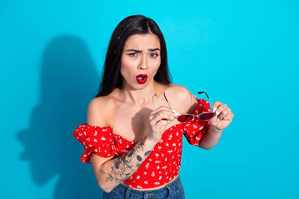 istock Charming young woman holding sunglasses with a surprised expression against a vibrant blue background in a casual red floral top 2212521328