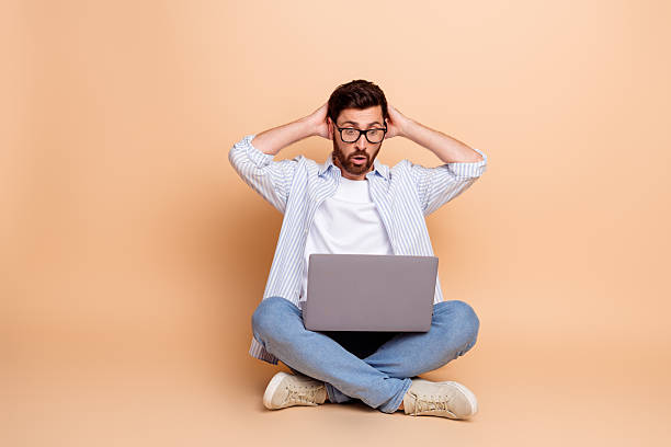 istock Young brunet man with glasses in casual style using laptop while gesturing surprise on beige studio background 2212523976