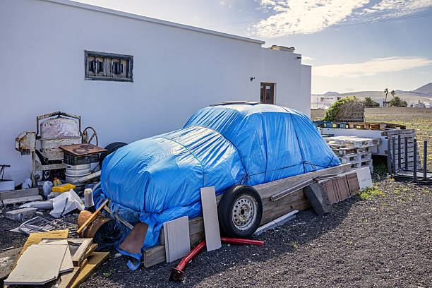 Nazaret, Lanzarote, Canary Islands, Spain - March 16th 2025: This striking urban photograph captures a car concealed beneath a blue tarpaulin, parked amidst scattered objects in what appears to be a storage or junkyard setting. Wooden pallets, a discarded tire, and miscellaneous debris surround the covered vehicle, creating a contrast between preservation and neglect. Set against the backdrop of a simple white building, the scene evokes themes of time, transience, and functionality in Lanzarote’s rural environment.