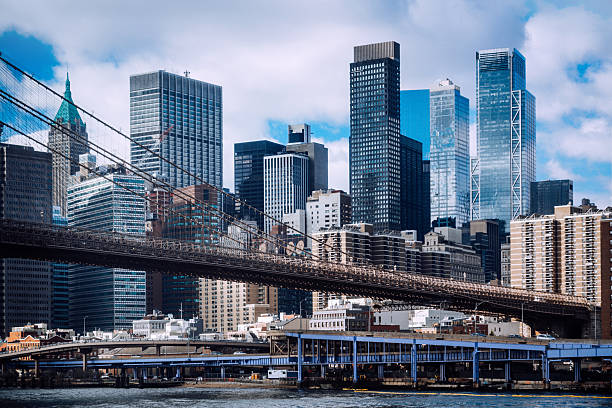 View of the iconic Brooklyn Bridge spanning the East River with the modern Manhattan skyline in the background, under a vibrant blue sky with scattered white clouds, showcasing urban architecture.