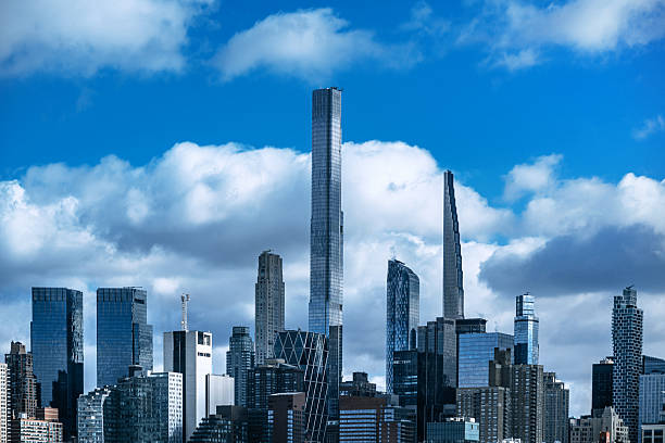 The image captures the Manhattan skyline featuring sleek, modern skyscrapers against a backdrop of a vibrant blue sky with scattered white clouds.