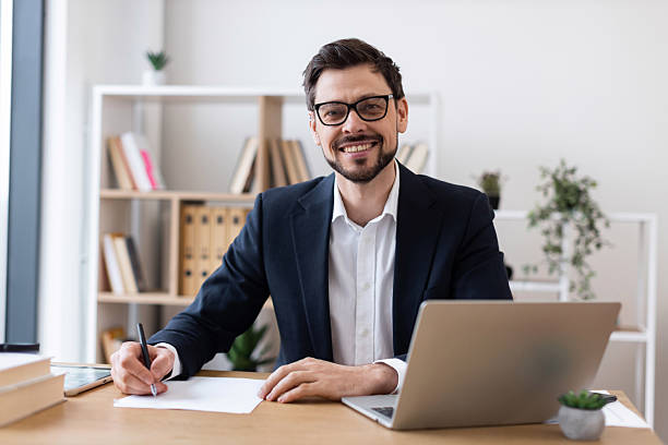 istock Smiling businessman writing notes at desk with laptop in modern office 2212527760
