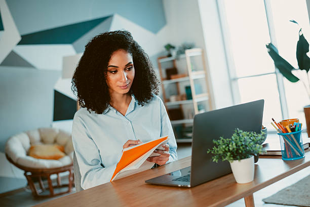 istock Young woman with curly hair taking notes while working on laptop in cozy modern home office during daytime 2212528330