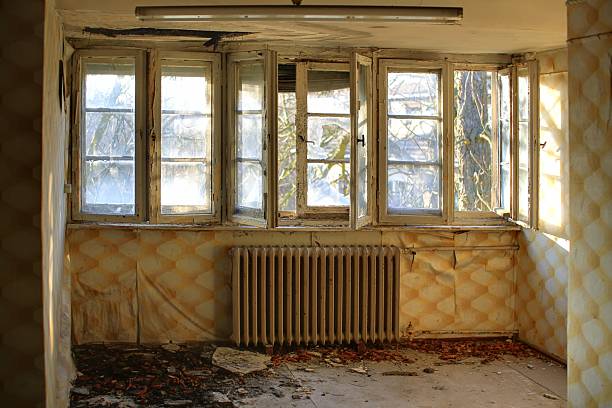 Broken old windows in a decayed room with peeling wallpaper and a rusted radiator below.