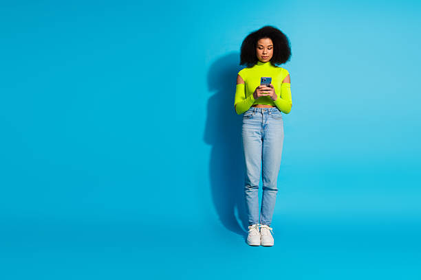 istock Young woman with neon green top using smartphone while standing against solid blue background in casual stylish attire 2212531747