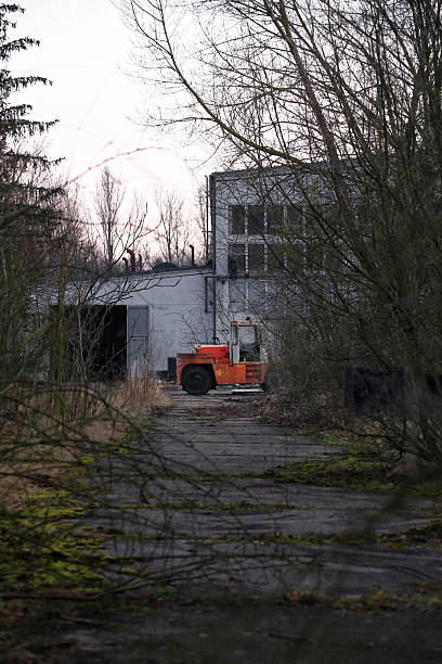 Rusty, orange forklift on overgrown yard in front of desolate industrial building.