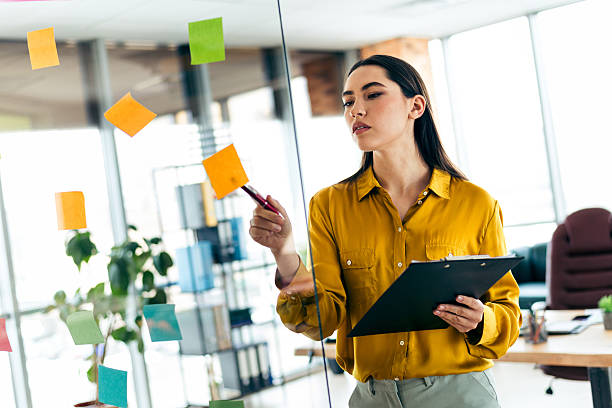 istock Young businesswoman organizing tasks with sticky notes on a transparent board, showcasing a modern workplace environment 2212532076