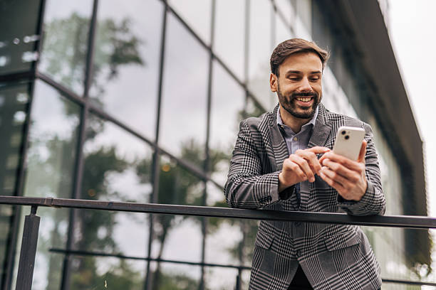 istock Smiling businessman using smartphone leaning on railing in front of office building 2212535119