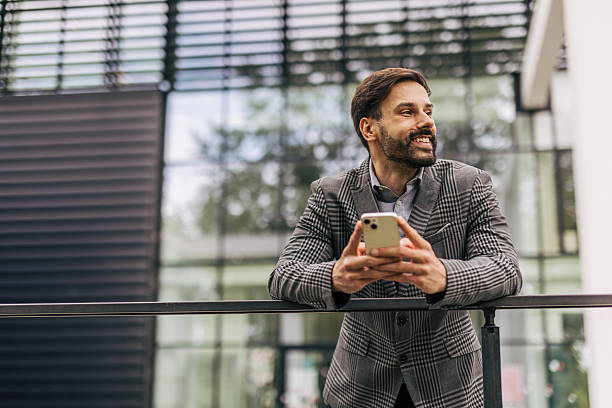 istock Smiling businessman leaning on railing and using smartphone outside office 2212535784