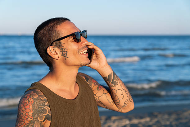 Happy tattooed young man talking on the phone at the beach