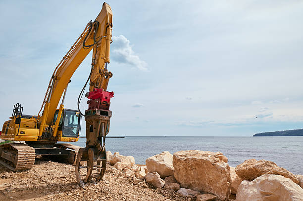 istock Yellow tracked excavator with hydraulic claw attachment working on a rocky coastal construction site near the sea under a partly cloudy sky 2212543591