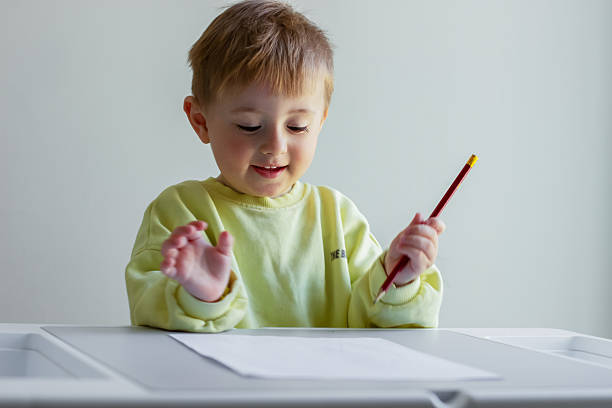 Cheerful little boy in a yellow sweater draws with colored pencils on paper at a table