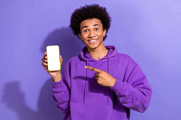 istock Portrait of optimistic young millennial guy hipster pointing finger phone display empty space ad web banner isolated on violet background 2212551168