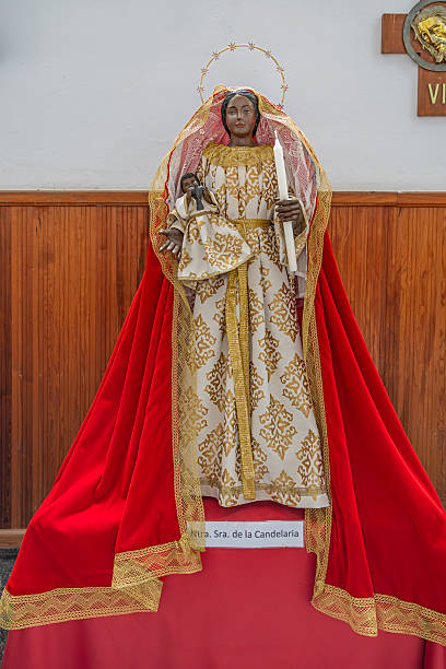 Nuestra Senora de la Encarnacion, Hara, Lanzarote, Canary Islands, Spain - March 13th 2025:  This striking religious picture captures the revered statue of Our Lady of Candelaria, displayed within the church of Nuestra Señora de la Encarnación. The saint is adorned in an elegant white and gold-patterned robe, complemented by a flowing red cape with golden trim. Holding a candle in one hand and a smaller figure in the other, she symbolizes devotion and spiritual guidance. A radiant halo encircles her head, enhancing the solemn yet majestic presence of the statue. The warm wooden paneling and religious inscriptions in the background further emphasize the sacred atmosphere of this revered site.