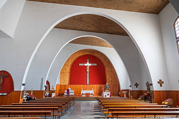 Nuestra Senora de la Encarnacion, Hara, Lanzarote, Canary Islands, Spain - March 13th 2025:  This photograph captures the interior of Nuestra Señora de la Encarnación in Haría, Lanzarote, with its elegant altar as the focal point. The altar is adorned with a large crucifix featuring Jesus Christ, set against a deep red backdrop that enhances the spiritual ambiance. The church’s modern architectural design features white arches and wooden elements, creating a serene and welcoming atmosphere. Rows of wooden pews lead toward the altar, inviting worshippers into a space of reflection and devotion. Religious statues and artifacts placed throughout the church further emphasize its sacred significance.