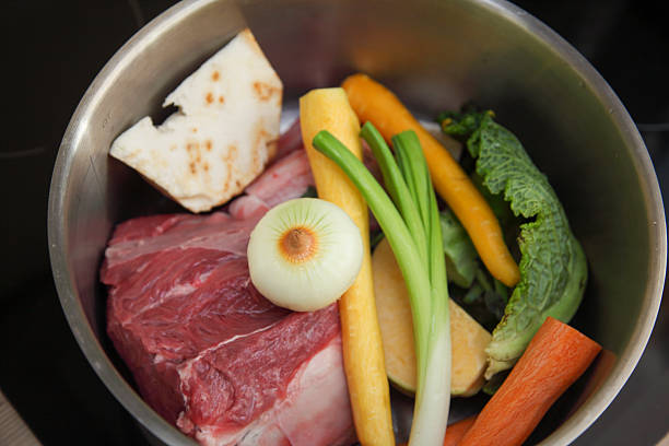 A top-down close-up of fresh ingredients for beef soup, including beef chunks, carrots, parsnip, celery root, leek, and possibly other vegetables, arranged in a stainless steel pot. Ready for cooking, this image illustrates the preparation of a hearty and traditional European soup. Ideal for illustrating concepts of soup ingredients, cooking, food preparation, healthy eating, and Mediterranean cuisine.