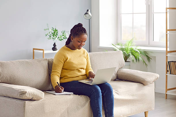 istock African American woman sitting on sofa, using laptop, studying online, and taking notes 2212561437