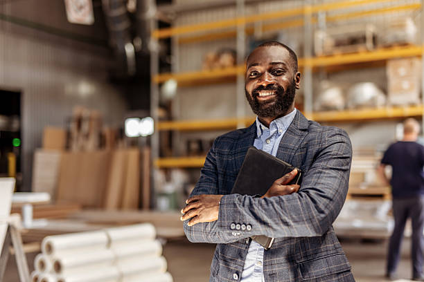 istock Smiling Businessman Standing in Warehouse with Rolled Blueprints and Shelves 2212562664