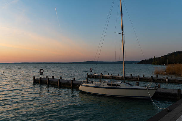 A sailboat is moored at a quiet pier during a peaceful sunrise, with soft pastel hues in the sky reflecting over the calm lake. The setting exudes stillness and solitude, perfect for themes of relaxation, retreat, or slow travel.