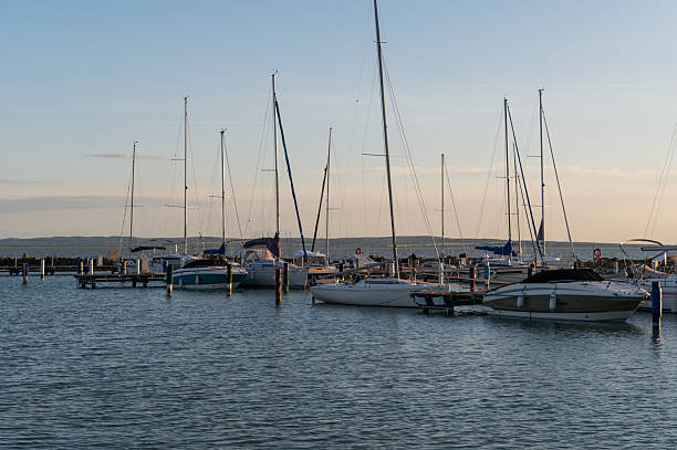 Several sailboats and motorboats are docked in a peaceful marina as the sun sets on the horizon. The calm water and warm evening light create a serene and inviting nautical scene.