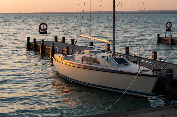 A small sailboat is tied to a wooden dock as the golden hour sun casts warm light across the water. The tranquil lake and calm atmosphere create a peaceful maritime setting.