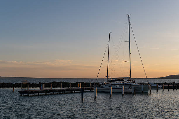 A sailboat is peacefully moored at a pier as the sun sets behind distant hills. The calm water and warm sky create a serene maritime scene at dusk.