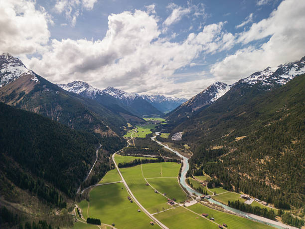 Aerial view of a picturesque Alpine valley featuring a winding river that flows through lush green meadows dotted with scattered farmhouses. The valley is framed by majestic, snow-covered mountain peaks and dense coniferous forests. The sky is partly cloudy, adding a dramatic yet peaceful atmosphere to the scene. This panoramic image evokes a sense of vastness, freedom, and untouched nature—perfect for themes such as nature, travel, sustainability, and adventure.