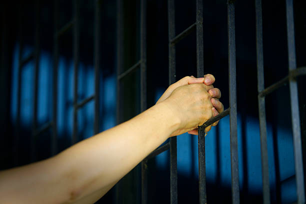 istock Hand Grasping Metal Bars of Prison Cell in Dimly Lit Environment 2212573835