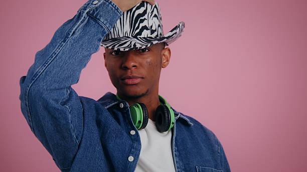 A young man showcases his unique style with a striking zebra-striped hat and denim jacket, perfectly blending urban fashion with playful charm in a lively pink backdrop.